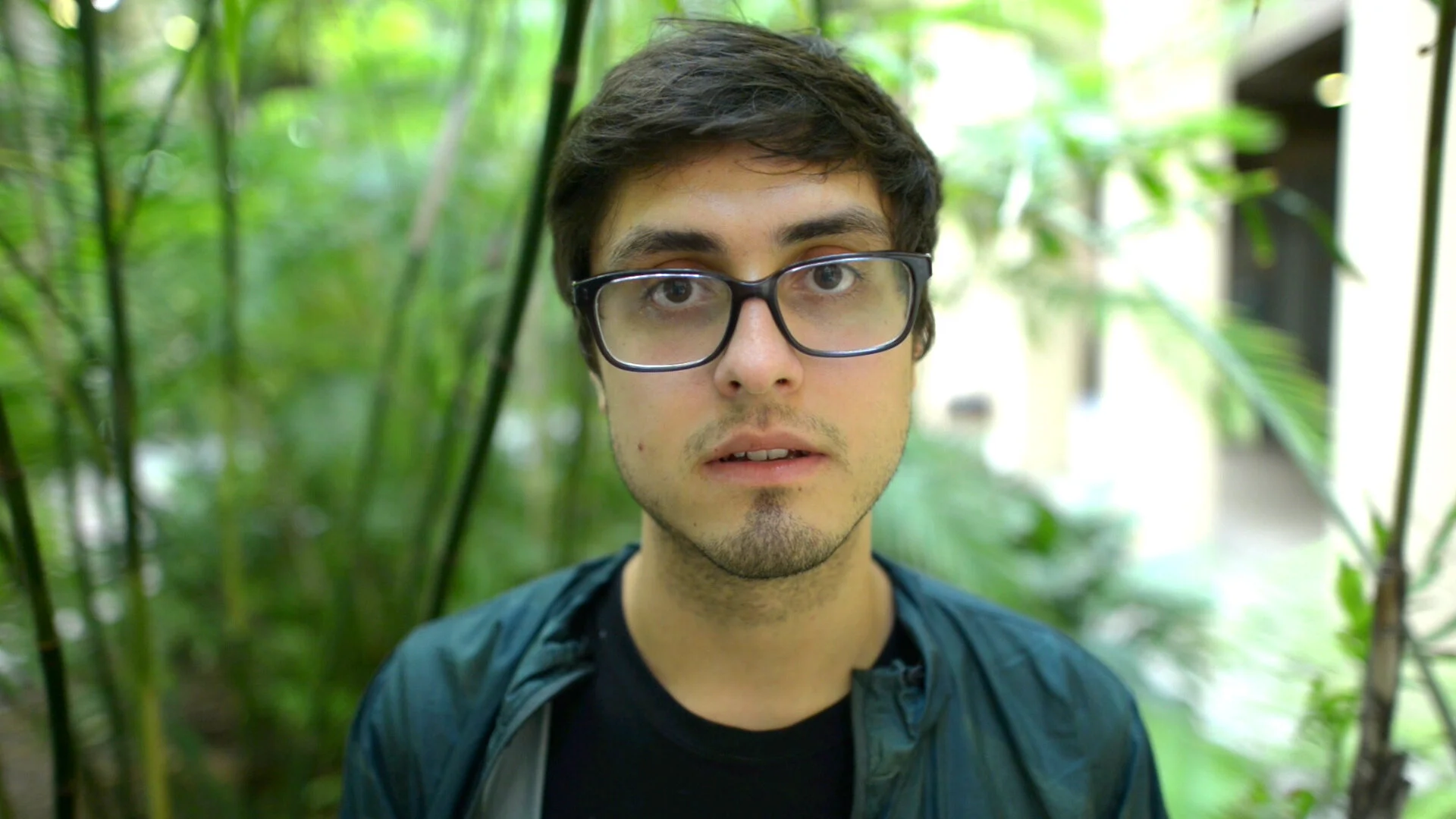 A young man with glasses and dark hair standing outdoors with green foliage in the background.