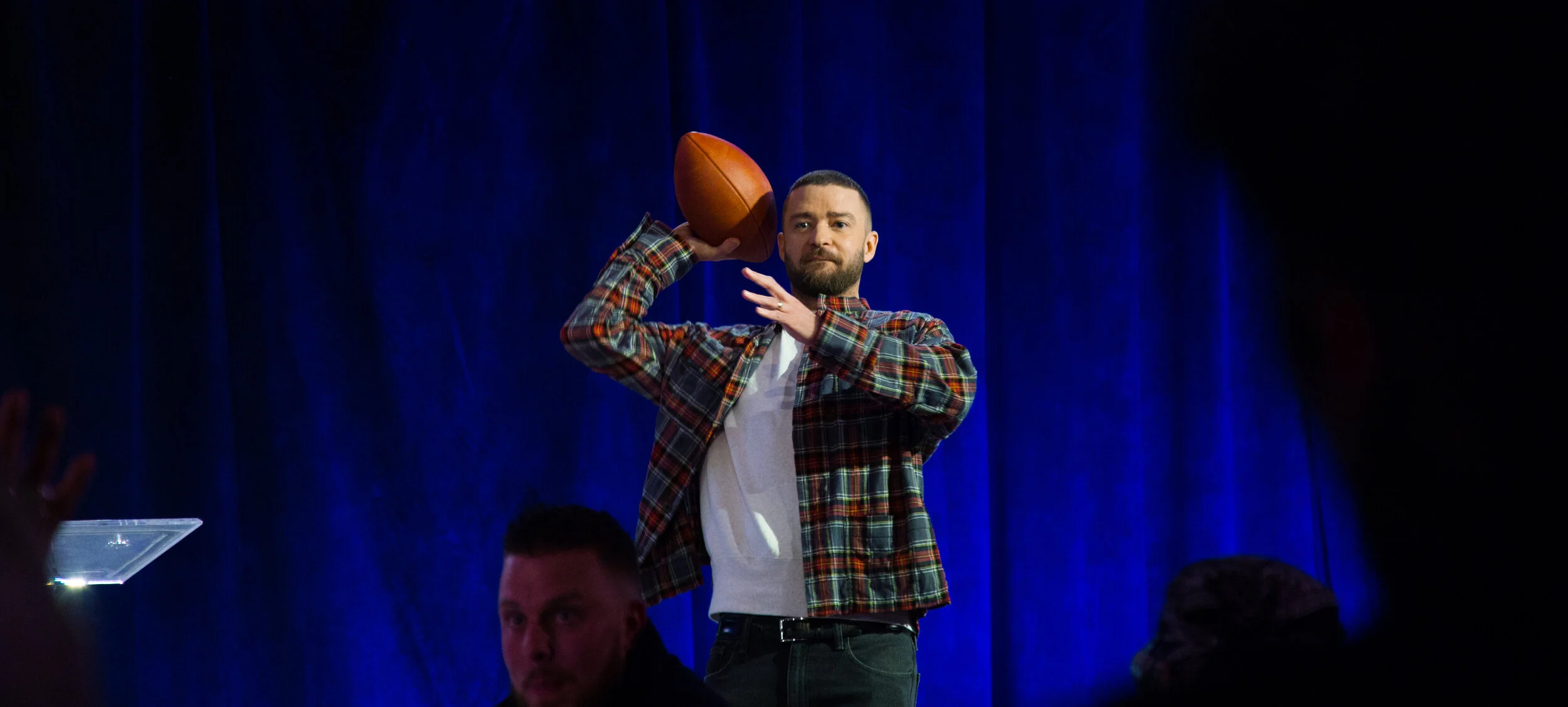 Justin Timberlake wearing a plaid shirt and a white T-shirt is holding a football in his right hand, raised to his ear, on a stage with a dark blue curtain backdrop at a Super Bowl LII press event.