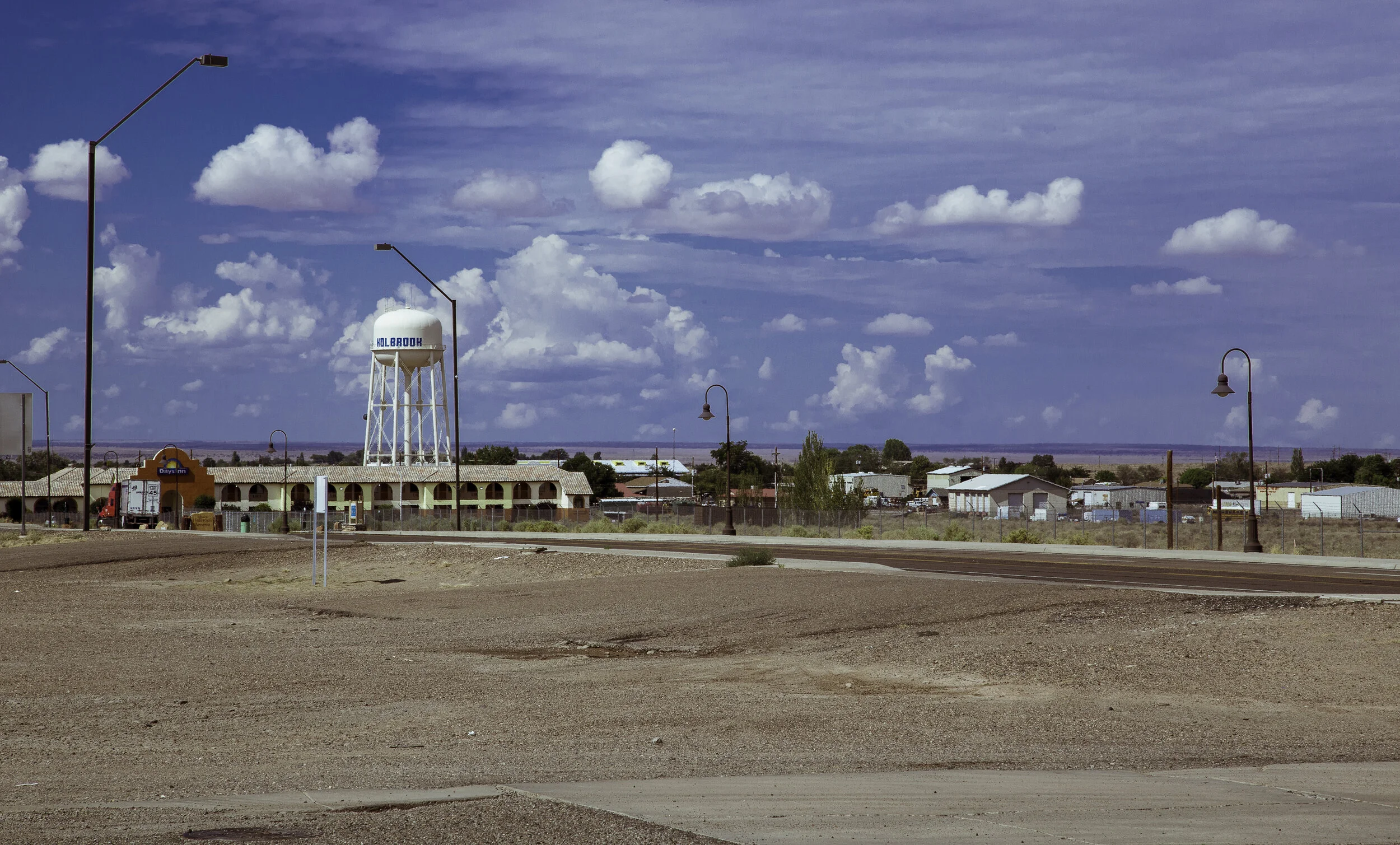 Wide view of a small town with a tall water tower labeled 'Holabook', streetlights, and a row of buildings under a partly cloudy sky, with a dirt foreground and distant horizon.