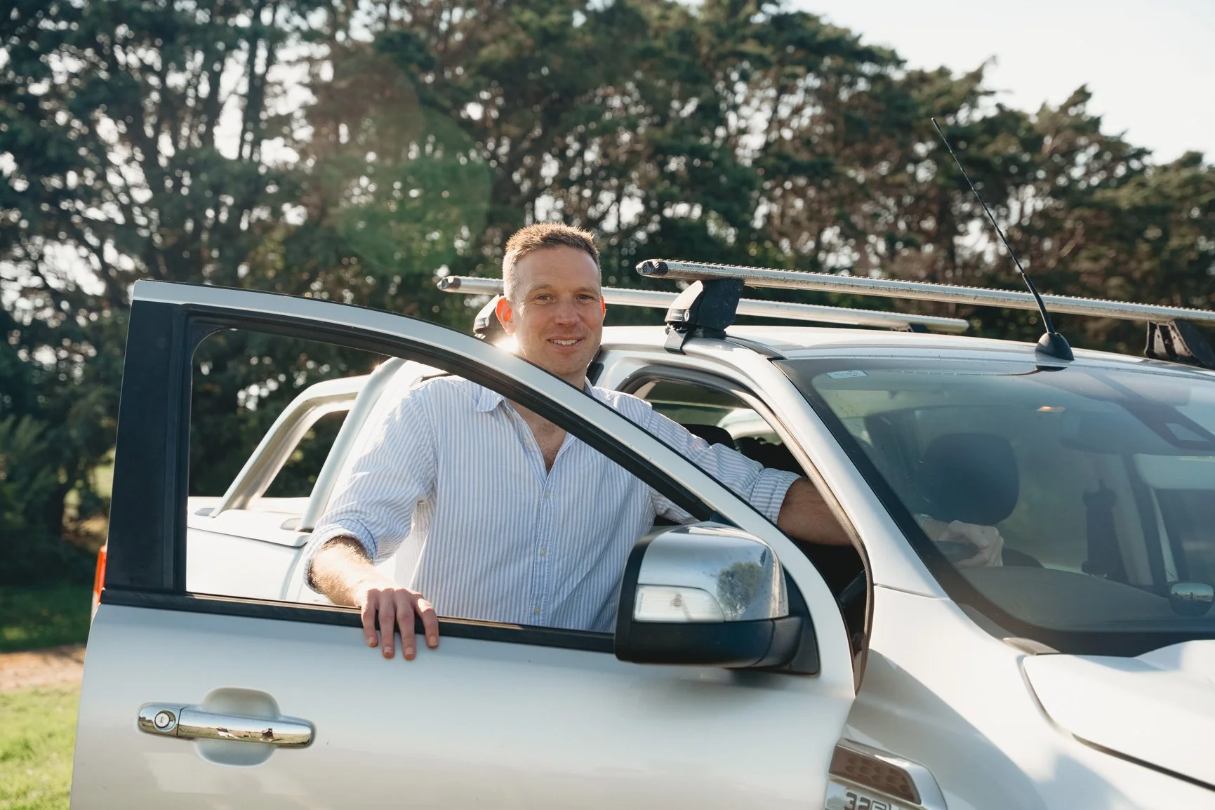 A man with short hair and a light blue striped shirt leaning out of the driver's side of a white pickup truck, smiling, with trees in the background on a sunny day.