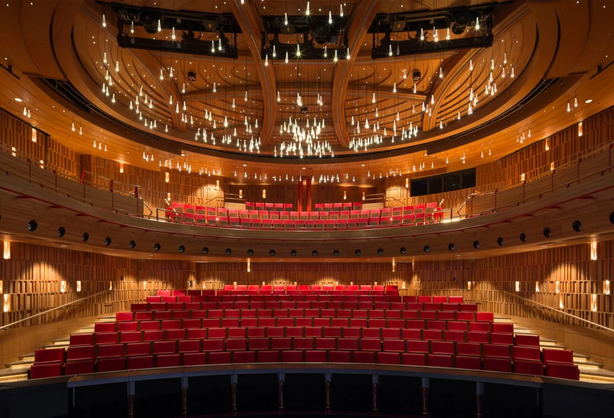 Royal Academy of Music. Susie Sainsbury Theatre. Interior of a modern concert hall with wooden walls, red seats, and a ceiling decorated with hanging lights.