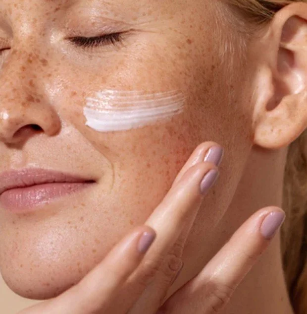 Close-up of a woman with freckles applying moisturiser on her cheek, using her finger.