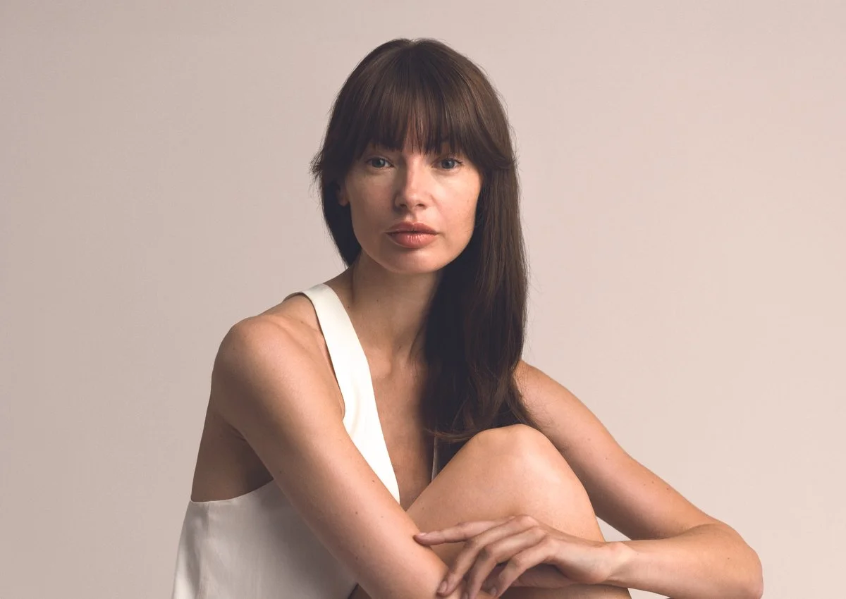 A woman with long brown hair and bangs sitting against a plain light-coloured background, wearing a white sleeveless top, with her knees drawn up and her arms wrapped around her leg.