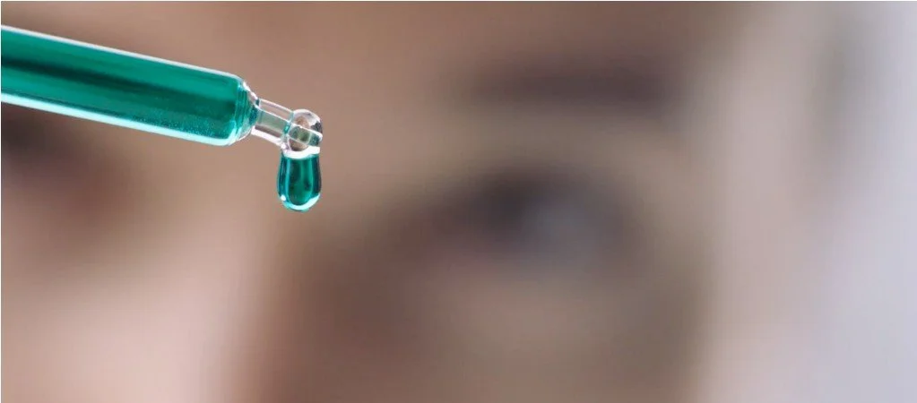 A close-up of a dropper with a teal-colored liquid about to release a droplet, with a blurred person's face in the background.