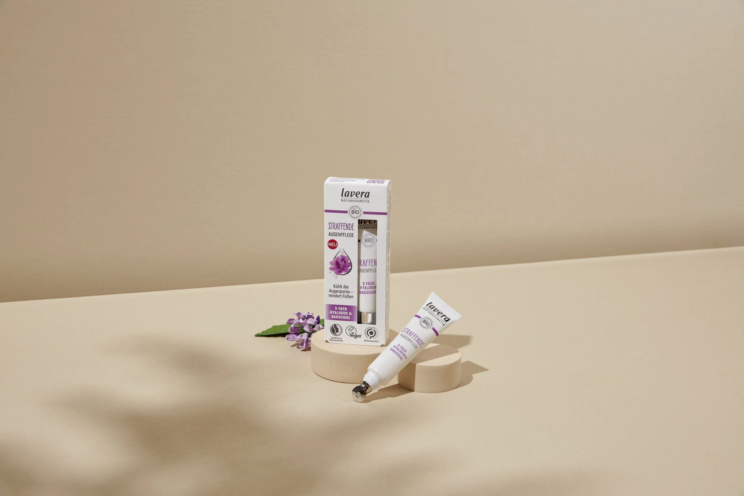 lavera Firming Eye Cream on a beige surface, decorated with purple flowers and green leaves.