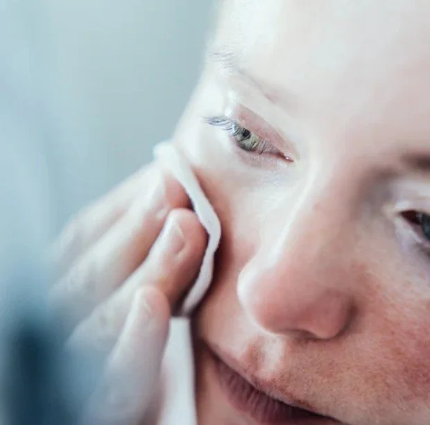 Close-up of a woman gently wiping her face with a cotton pad, focusing on her eye and cheek.