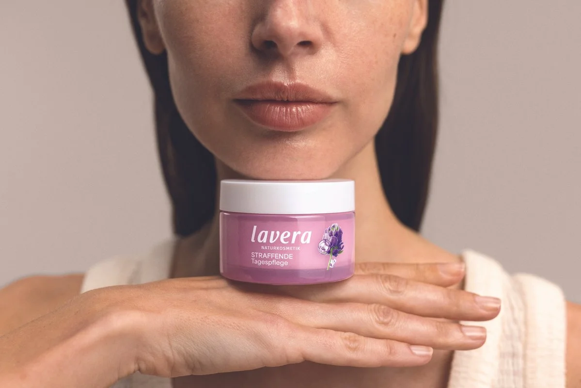 A woman with a jar of lavera Firming Day Cream near her chin, with her hand supporting the jar. The focus is on the jar and her lips, with her face partly visible against a neutral background.