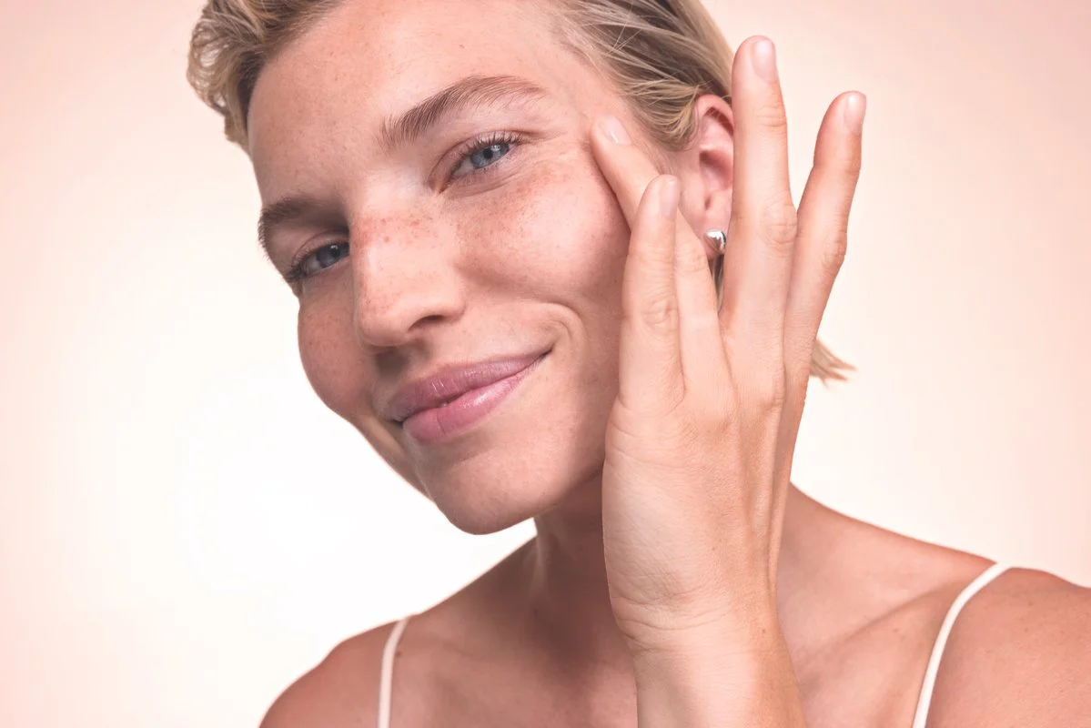 A young woman with short blonde hair, freckles, and blue eyes smiling softly, touching her face with her right hand.