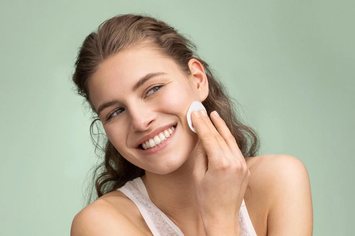 A young woman with long brown hair is smiling and applying skincare with a cotton pad on her cheek against a light green background.
