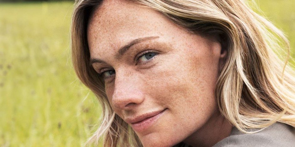 Close-up of a woman with blonde hair, freckles, and blue eyes smiling outdoors with a green background.