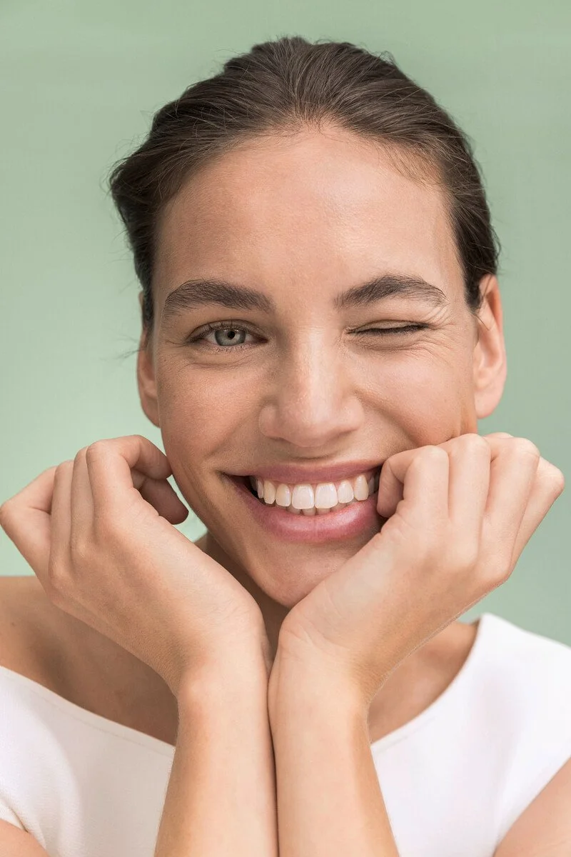 A woman smiling and winking, with her hands gently touching her face, against a light green background.