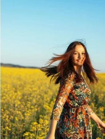 A woman with long hair standing in a field of yellow flowers under a clear blue sky.