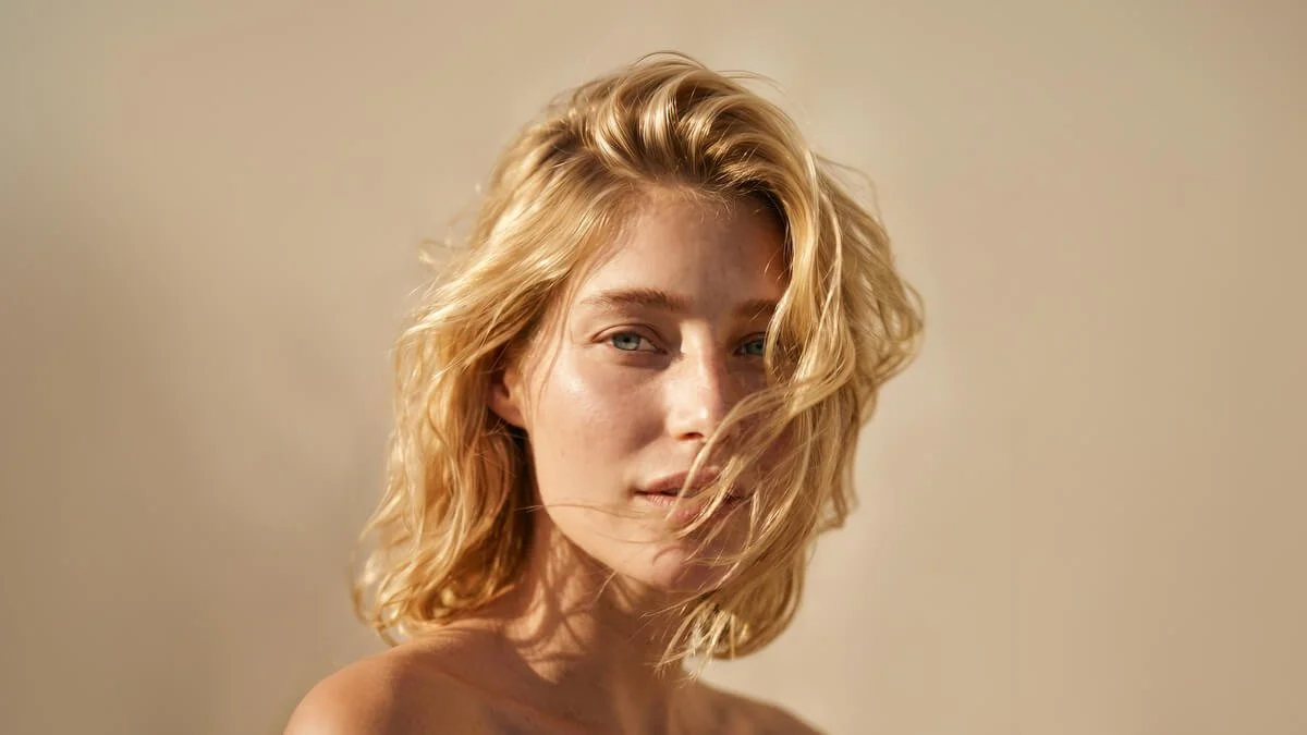Close-up of a young woman with wavy blonde hair and a neutral expression, with sunlight illuminating her face and hair against a plain, light-coloured background.