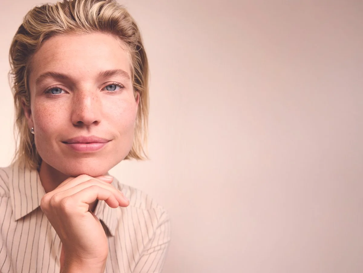 Close-up of a woman with blonde hair, blue eyes, and freckles, resting her chin on her hand against a plain beige background.