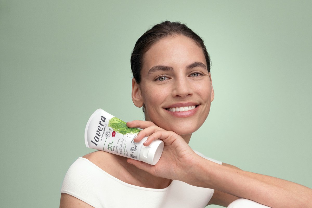 A smiling woman holding a tube of lavera Pure Beauty 3in1 Wash, Scrub Mask against a light green background.