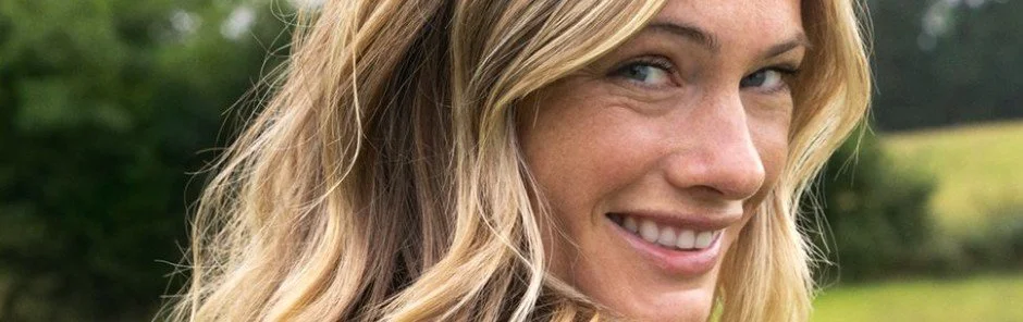 Close-up of a woman with blonde hair smiling outdoors, with trees and grass in the background.