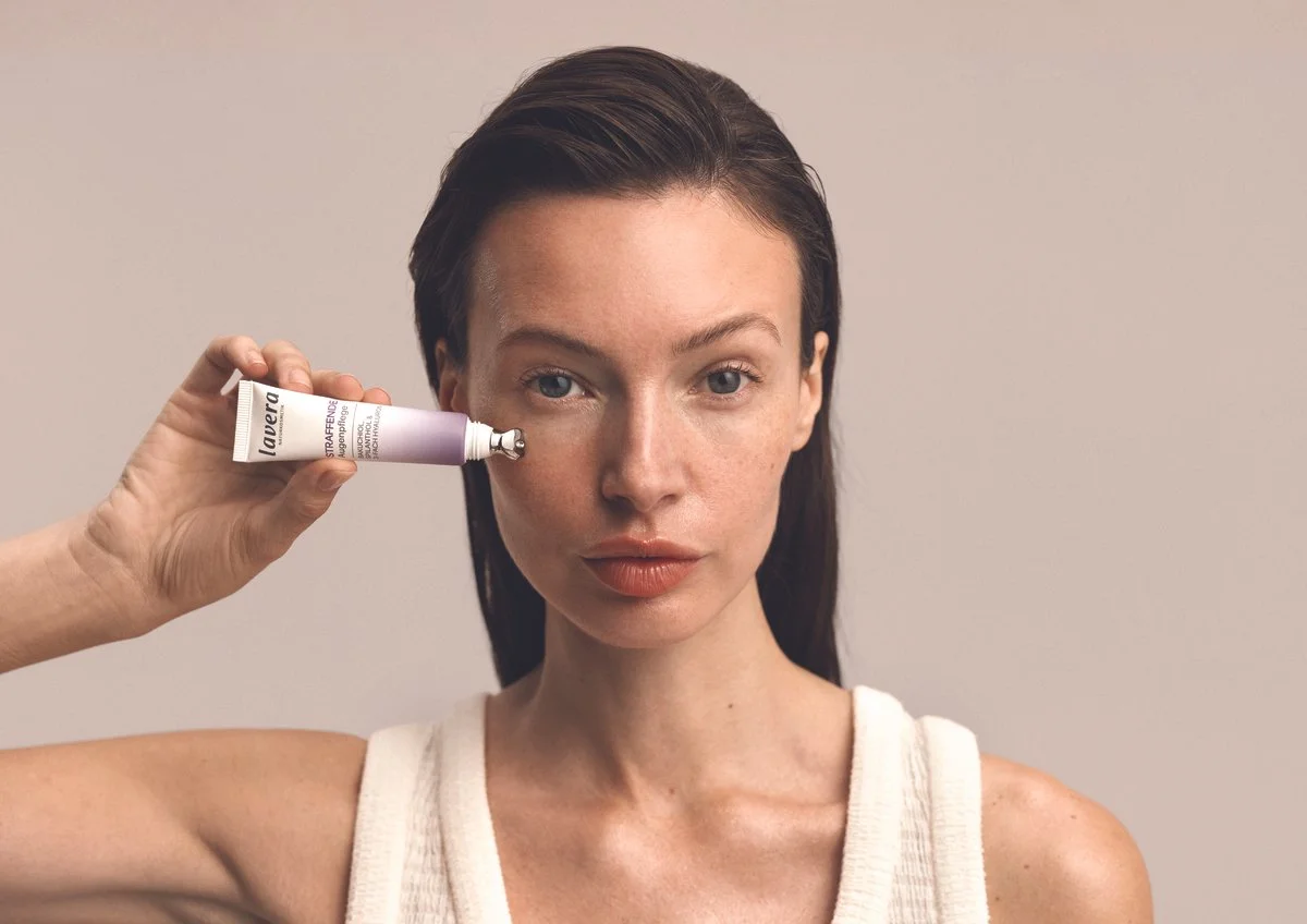 Woman with fair skin and dark hair applies skincare cream to her face using a tube from La Mer.
