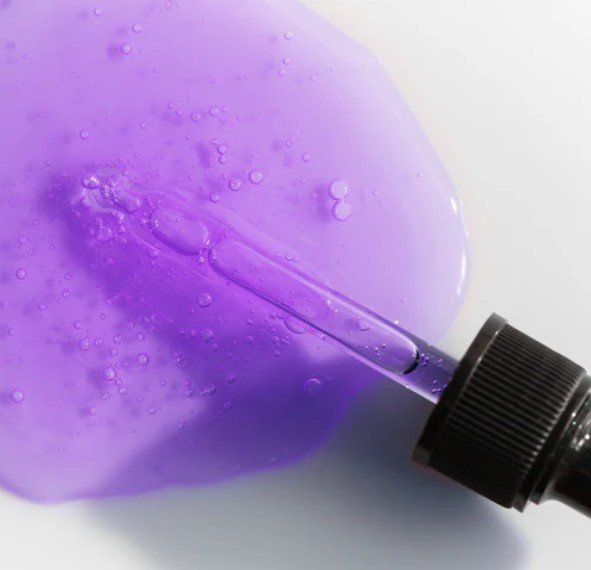Purple liquid with bubbles and a dropper laying on it on a white surface.