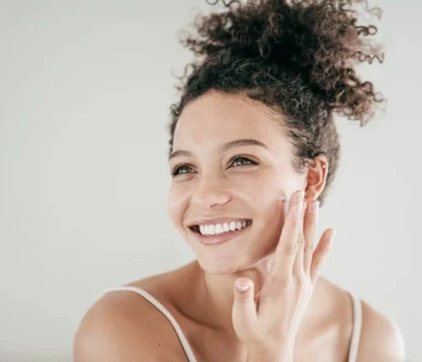 A young woman with curly hair smiling and touching her cheek.