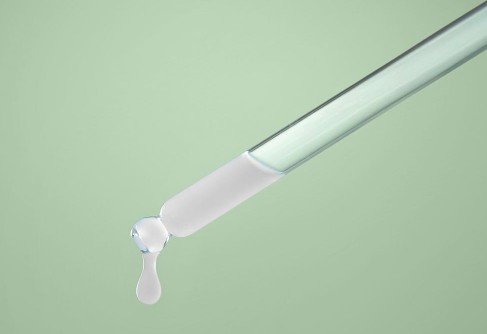 Close-up of a glass dropper with a droplet of liquid at the tip, against a light green background.