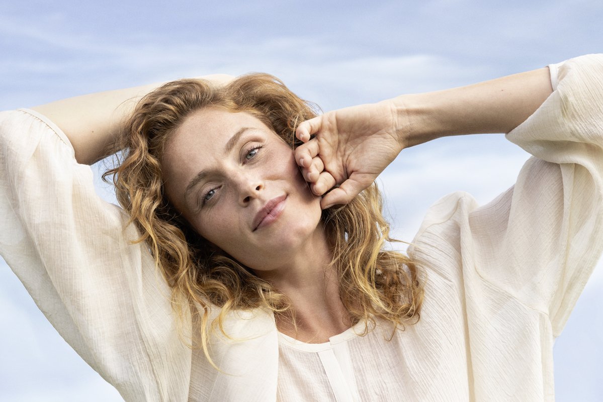 Woman with curly red hair relaxing outdoors, smiling softly with her eye closed, against a blue sky.