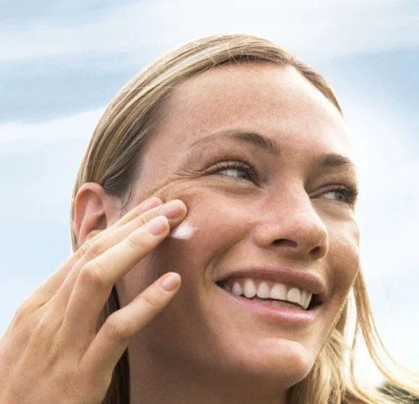A woman with blonde hair applying cream to her cheek outdoors, smiling with a cloudy sky in the background.