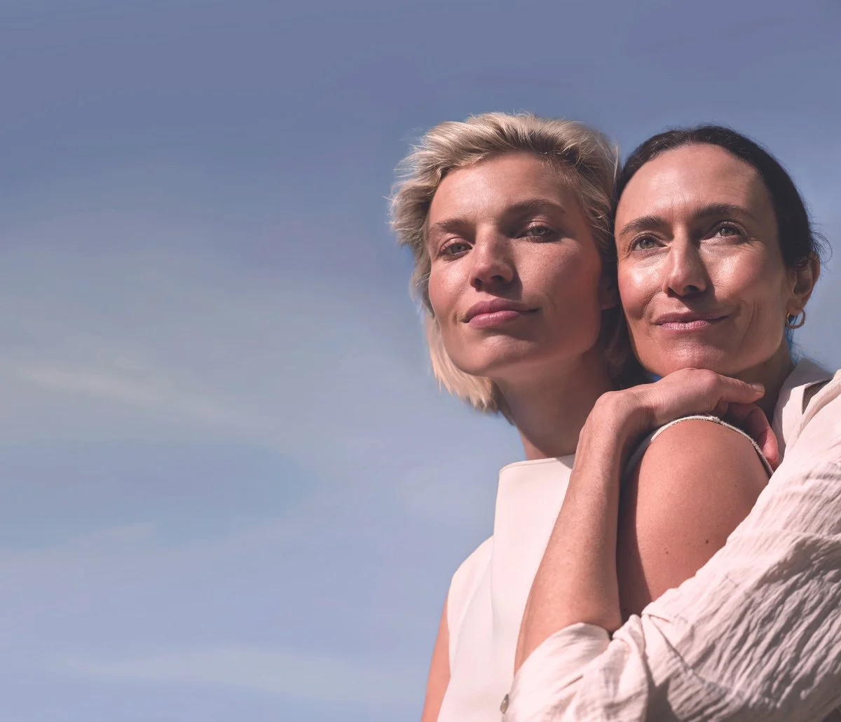 Two women embracing outdoors with a clear blue sky background, smiling softly.