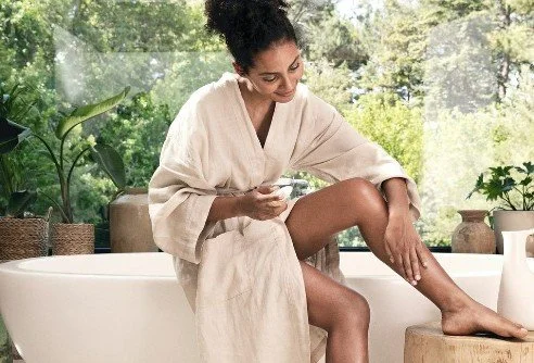 A woman in a beige robe sitting on a bathtub in a bright, green, plant-filled room, applying lotion or cream to her leg.