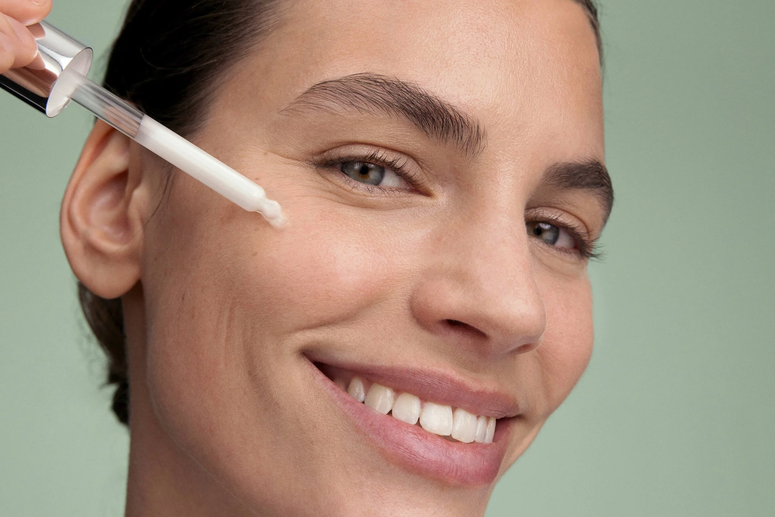 A woman with light skin and dark hair smiling while applying serum to her face.