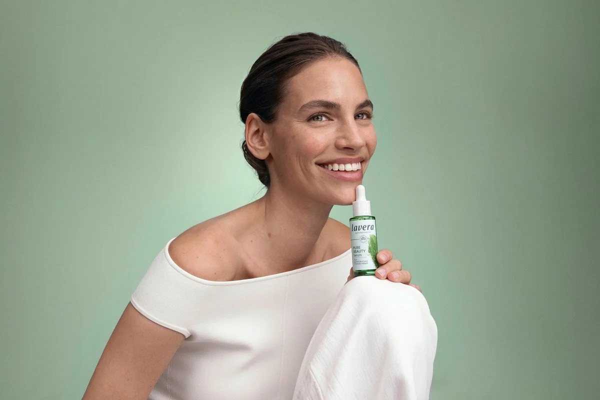 A smiling woman with short dark hair holding a bottle of lavera Pure Beauty Serum against a light green background.