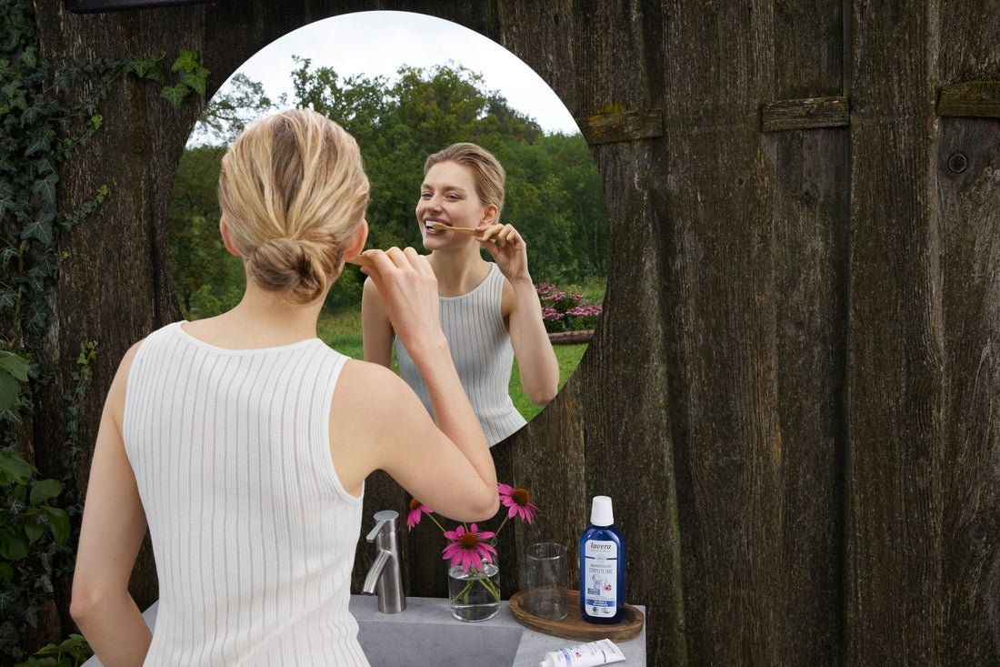 A woman brushing her teeth while looking into a mirror outside, surrounded by nature, with a bottle of mouthwash and other personal care items on the sink.