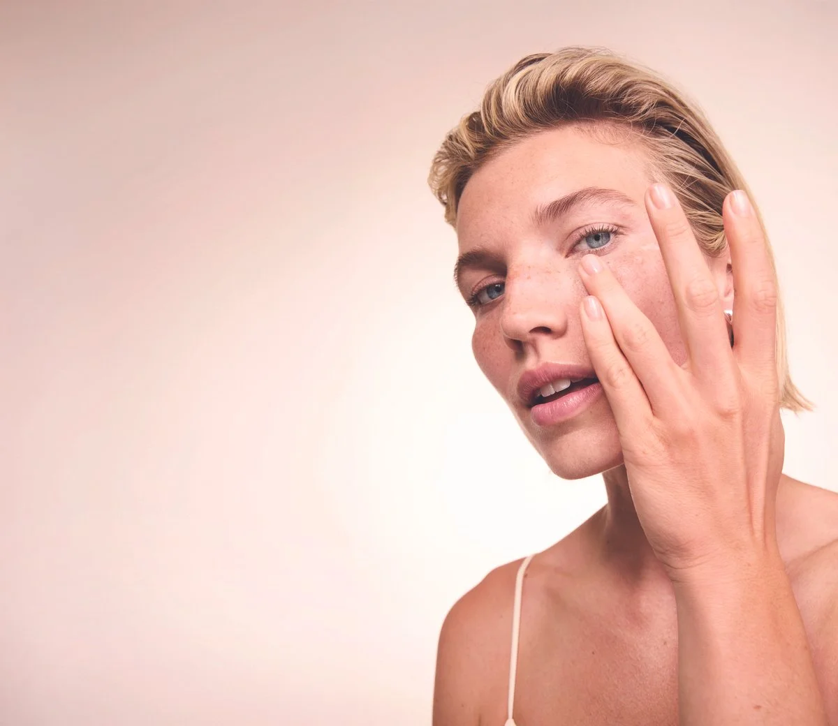 Close-up of a woman with short blonde hair touching her face with her right hand, showing clear skin and minimal makeup against a neutral background.