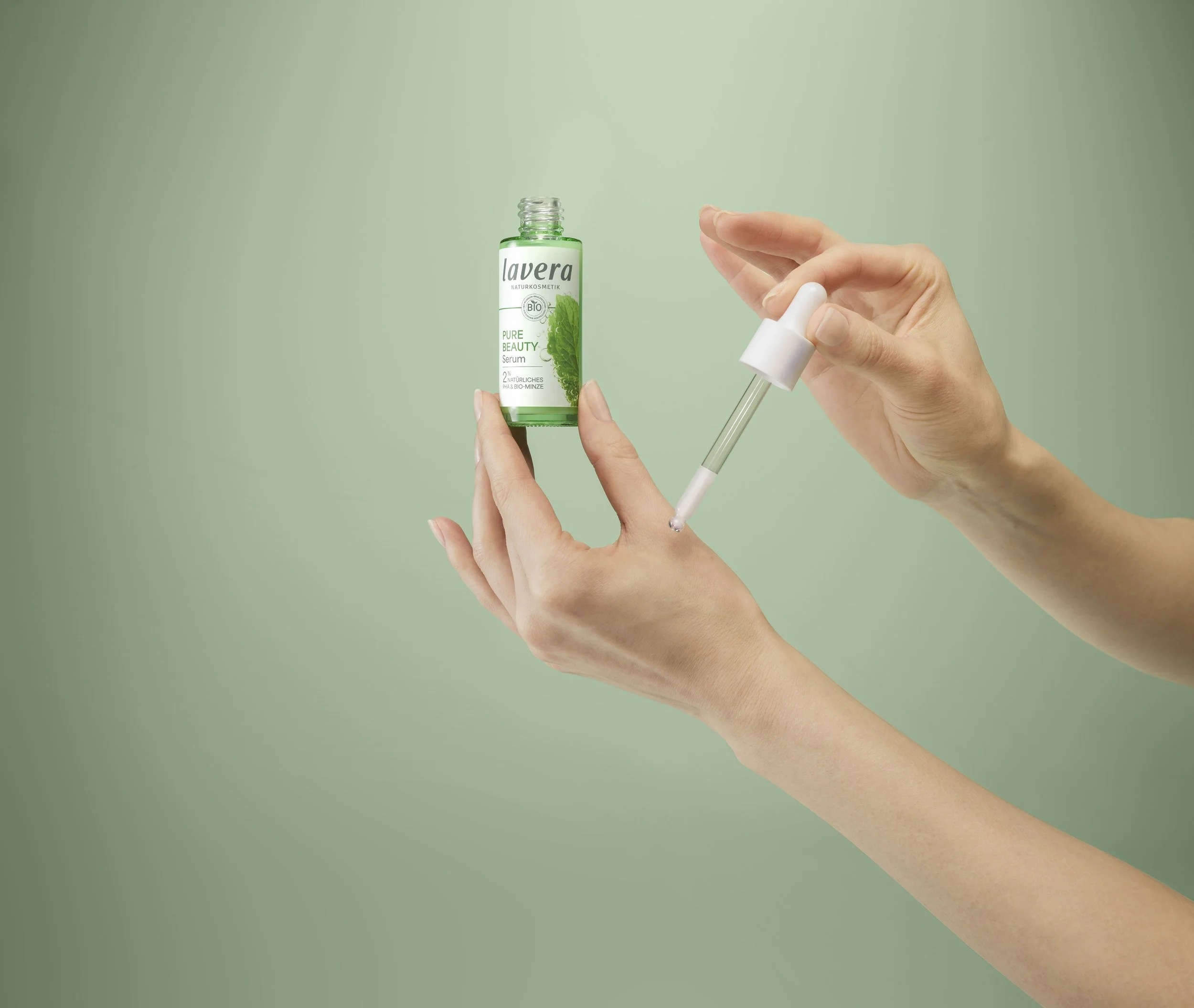 Hands holding a small green bottle of lavera Pure Beauty Serum and a dropper dispensing serum onto the hand.