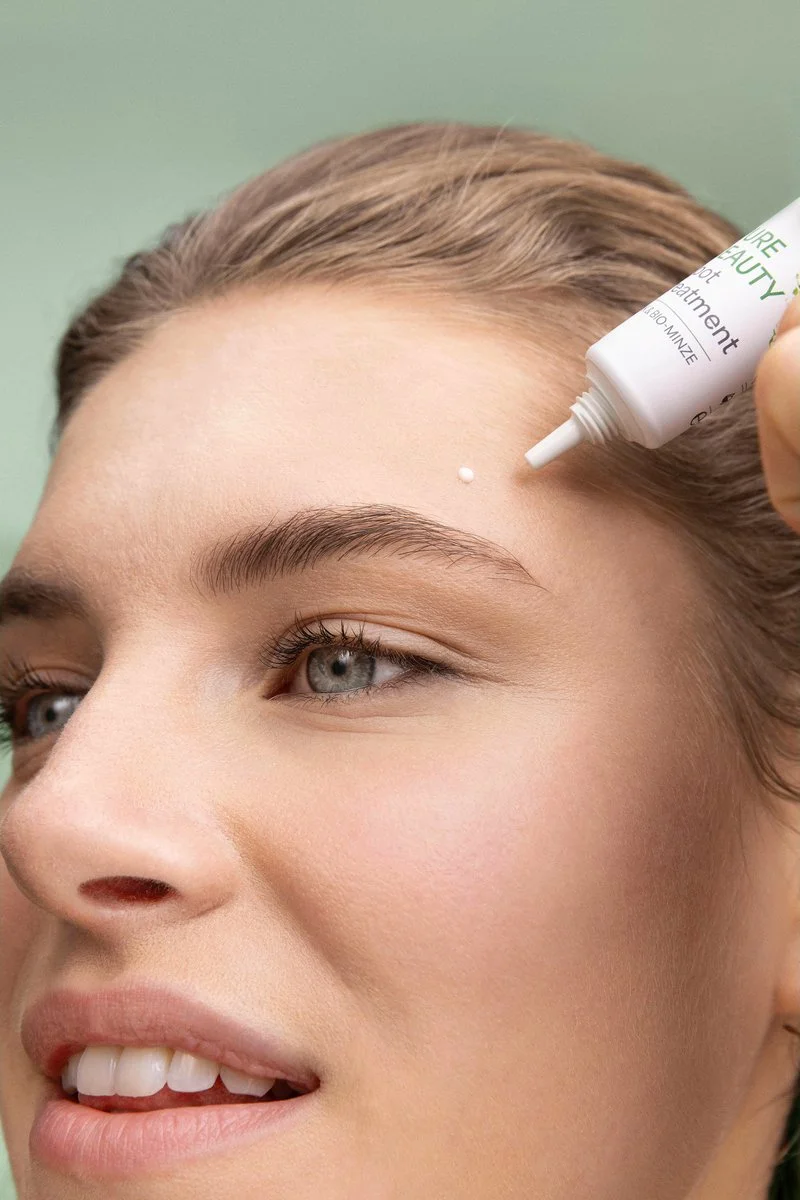 A woman with fair skin and light brown hair is applying a small amount of lavera Pure Beauty Spot Treatment product to her forehead.