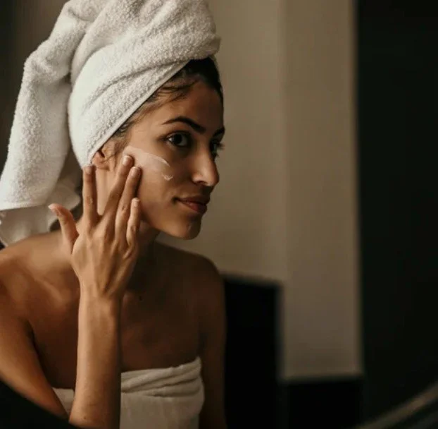 A woman wrapped in a towel with a towel turban on her head, applying skincare cream to her face in a bathroom.