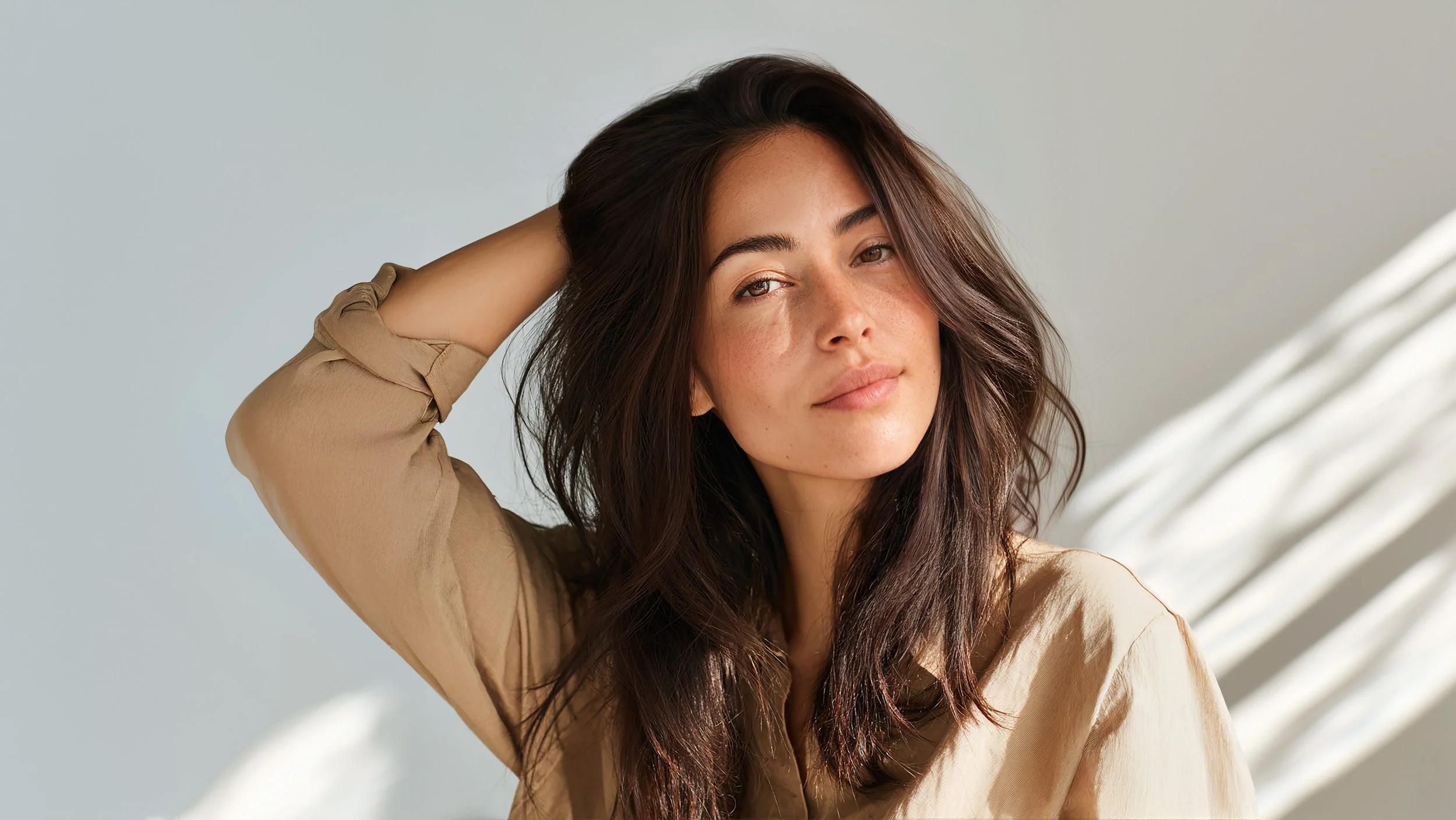 Close-up of a young woman with long brown hair, freckles, and a relaxed expression, wearing a beige top, with light coming through window blinds in the background.
