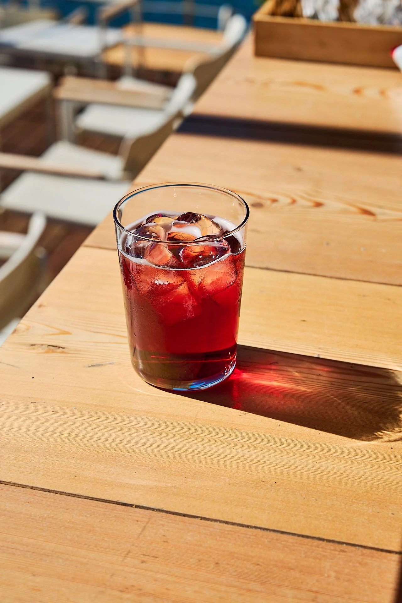 A glass of dark-colored iced beverage with ice cubes on a wooden table outdoors, with chairs visible in the background.