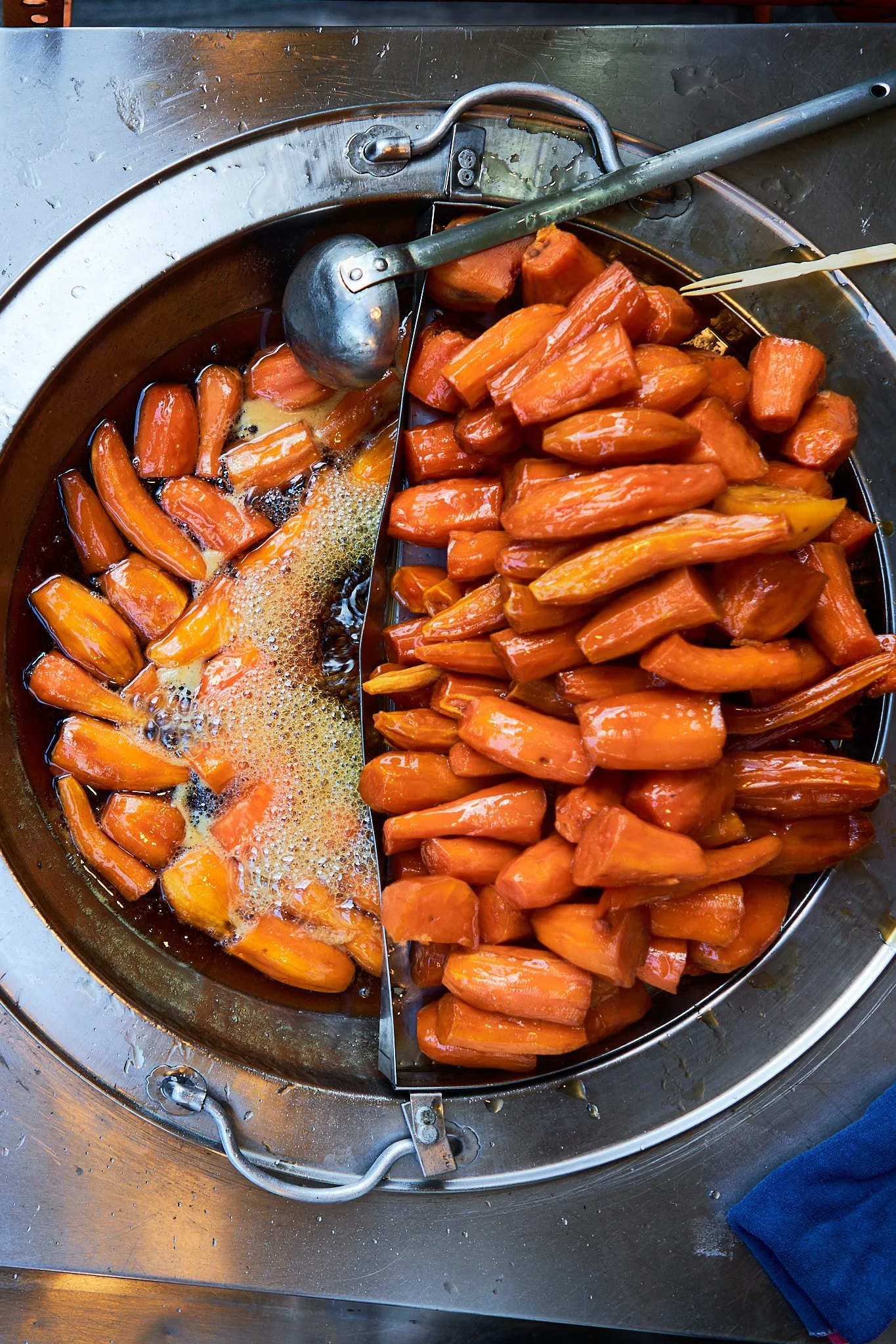 Carrots being cooked in a divided hot metal pan, with some carrots in a liquid and a serving spoon resting on the side.