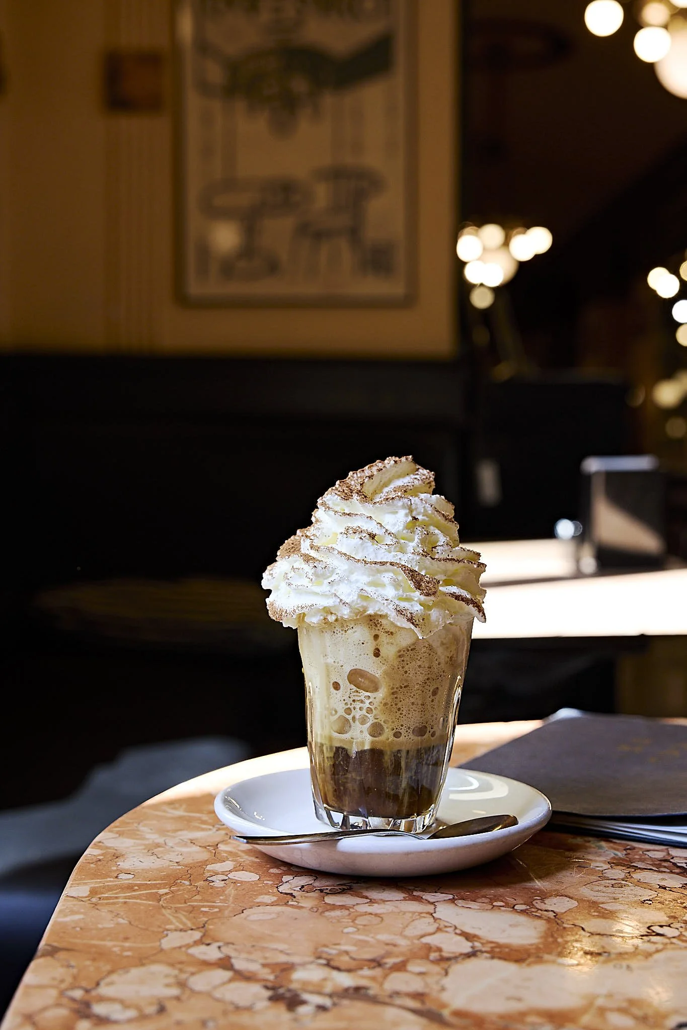 A glass of whipped cream-topped coffee drink on a saucer with a spoon, sitting on a marble table in a dimly lit café.