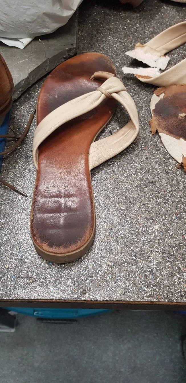 Verwüstetes, kaputtes Schuh, wahrscheinlich eine Sandale, mit Lederinnensohle und beschädigtem Riemen auf einem rauen Boden, neben einem zerbrochenen Stoffstück.