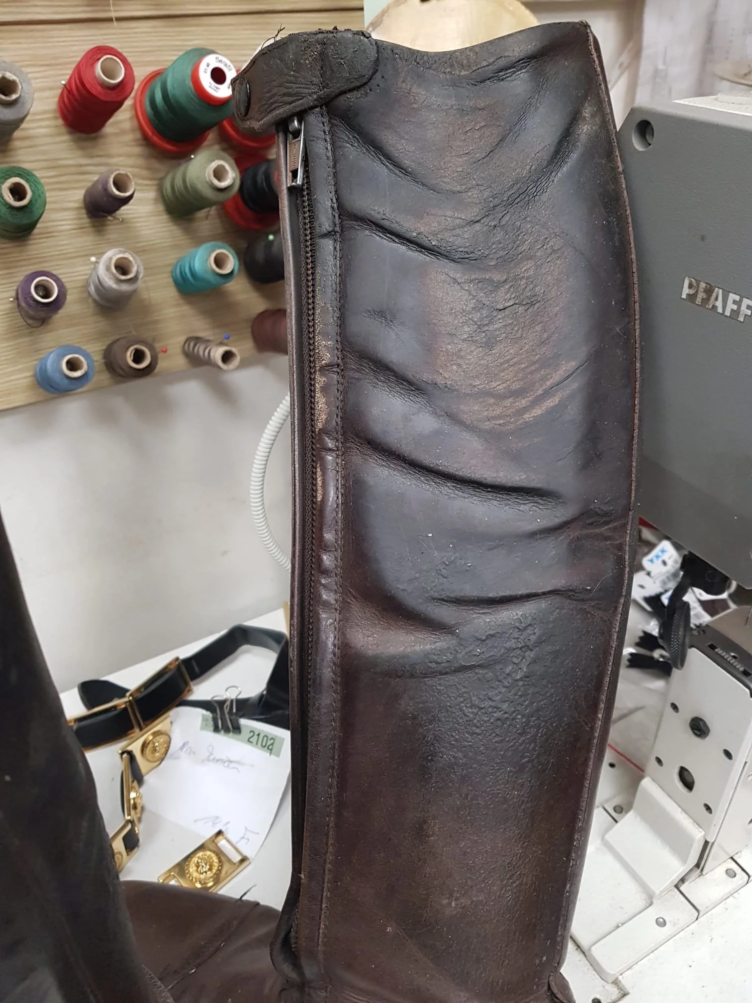 Ein brauner Lederriemen mit einer Reißverschluss, umgeben von verschiedenen Spulen mit buntem Garn auf einem Holztisch. Im Hintergrund sind Schmuckstücke sichtbar.