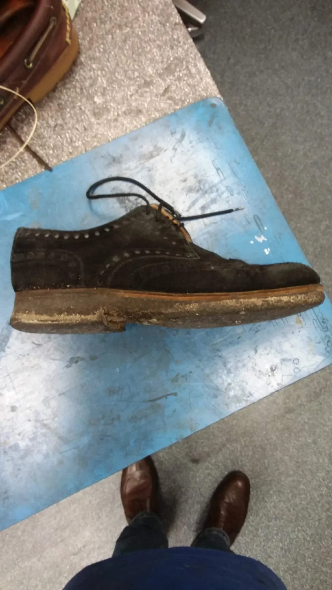 Eine angetragene schwarze Lederstiefel, die sich auf einem blauen Papier mit einem Schuhkarton im Hintergrund befinden, und man sieht die Füße und das Bein einer Person, die vor dem Bild steht.