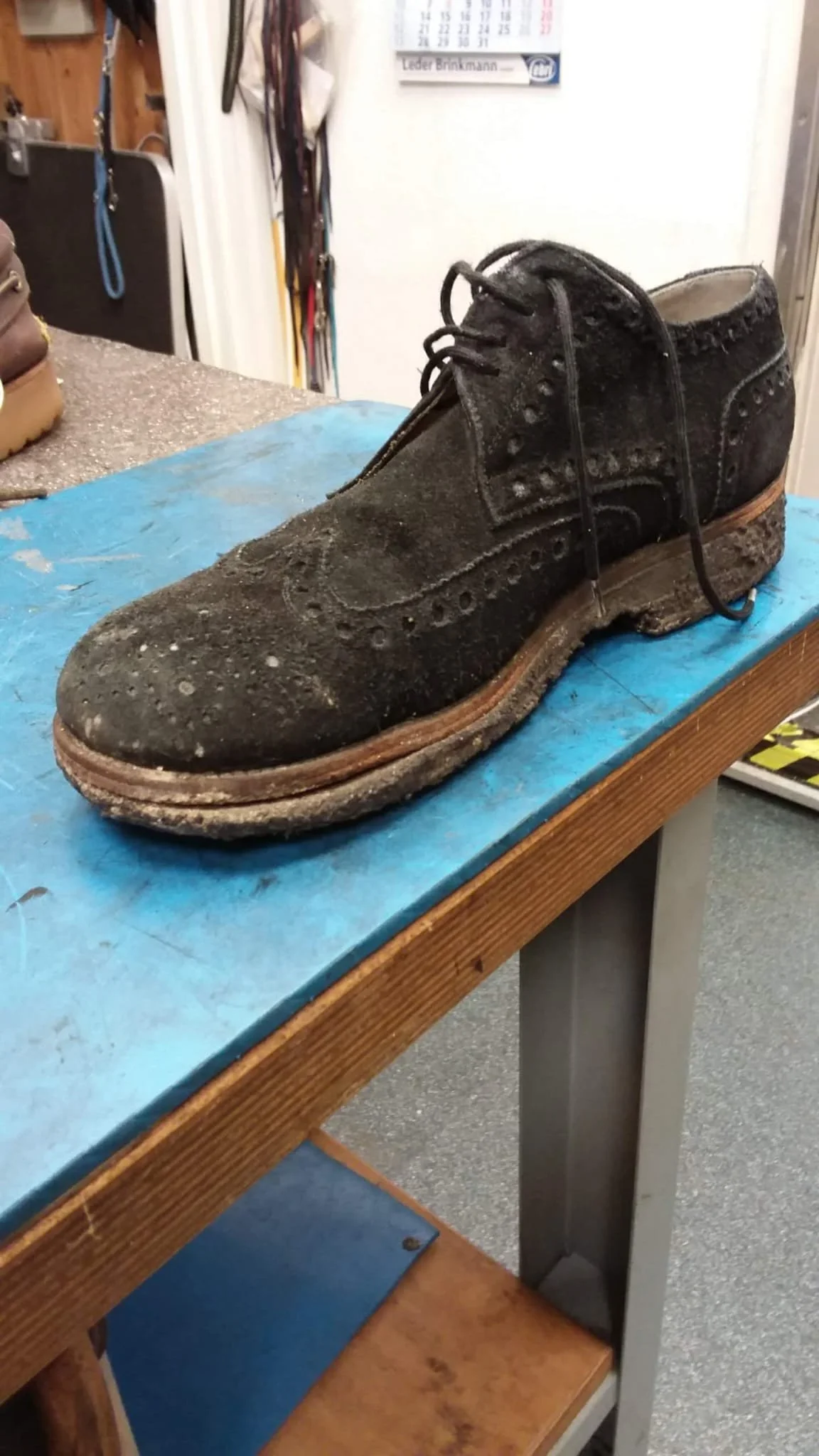 Ein alter, abgenutzter schwarzer Lederschuh mit zahlreichen Staub- und Schmutzspuren liegt auf einem blauen Tisch. Die Schuhe sind zerkratzt und tragen Anzeichen von Abnutzung.
