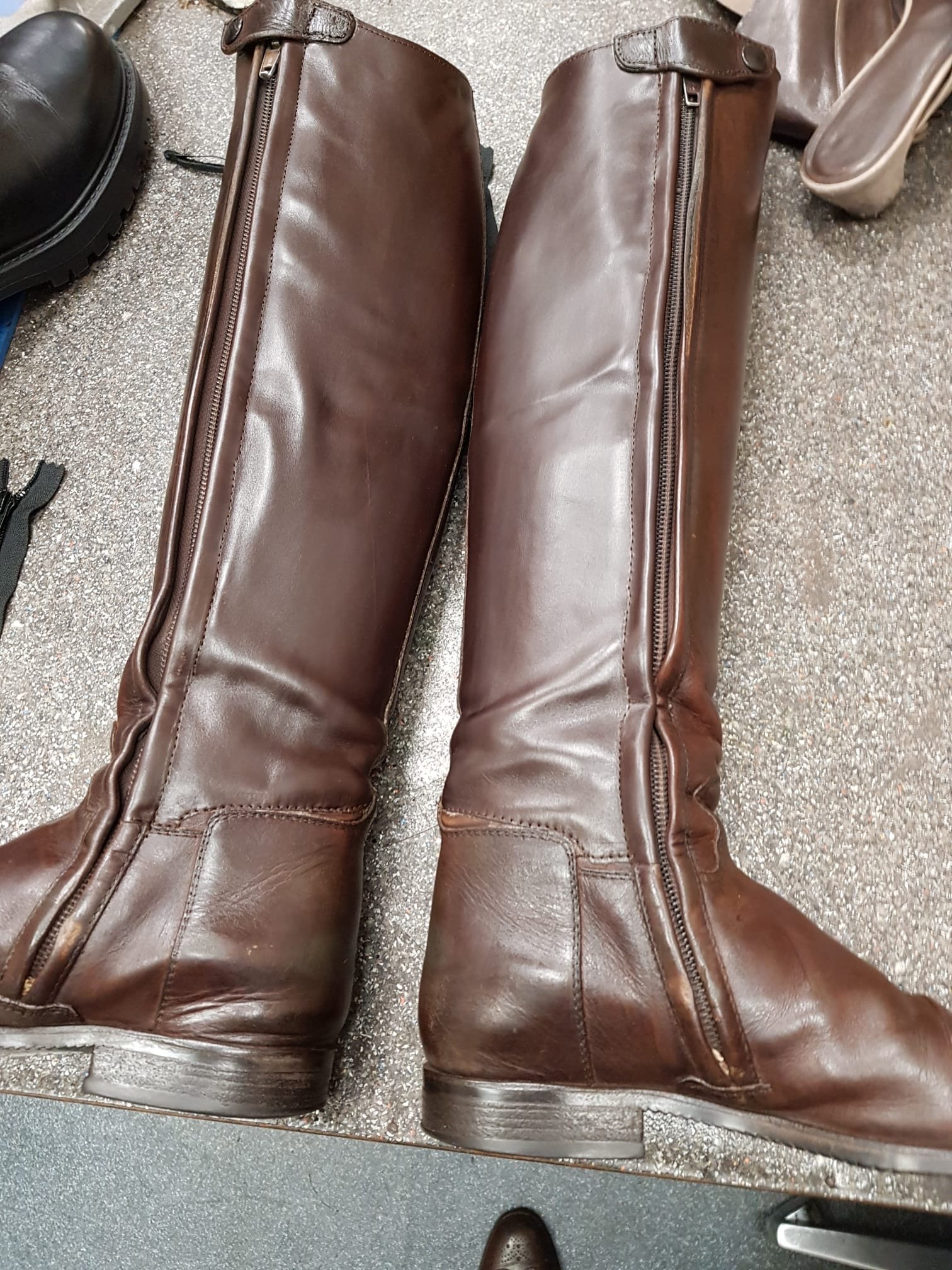 Braune, kniehohe Stiefel aus Leder mit Zippern an der Seite, getragen auf einer Oberfläche mit anderen Schuhen und Gegenständen, wahrscheinlich in einem Laden oder in einer Umkleidekabine.