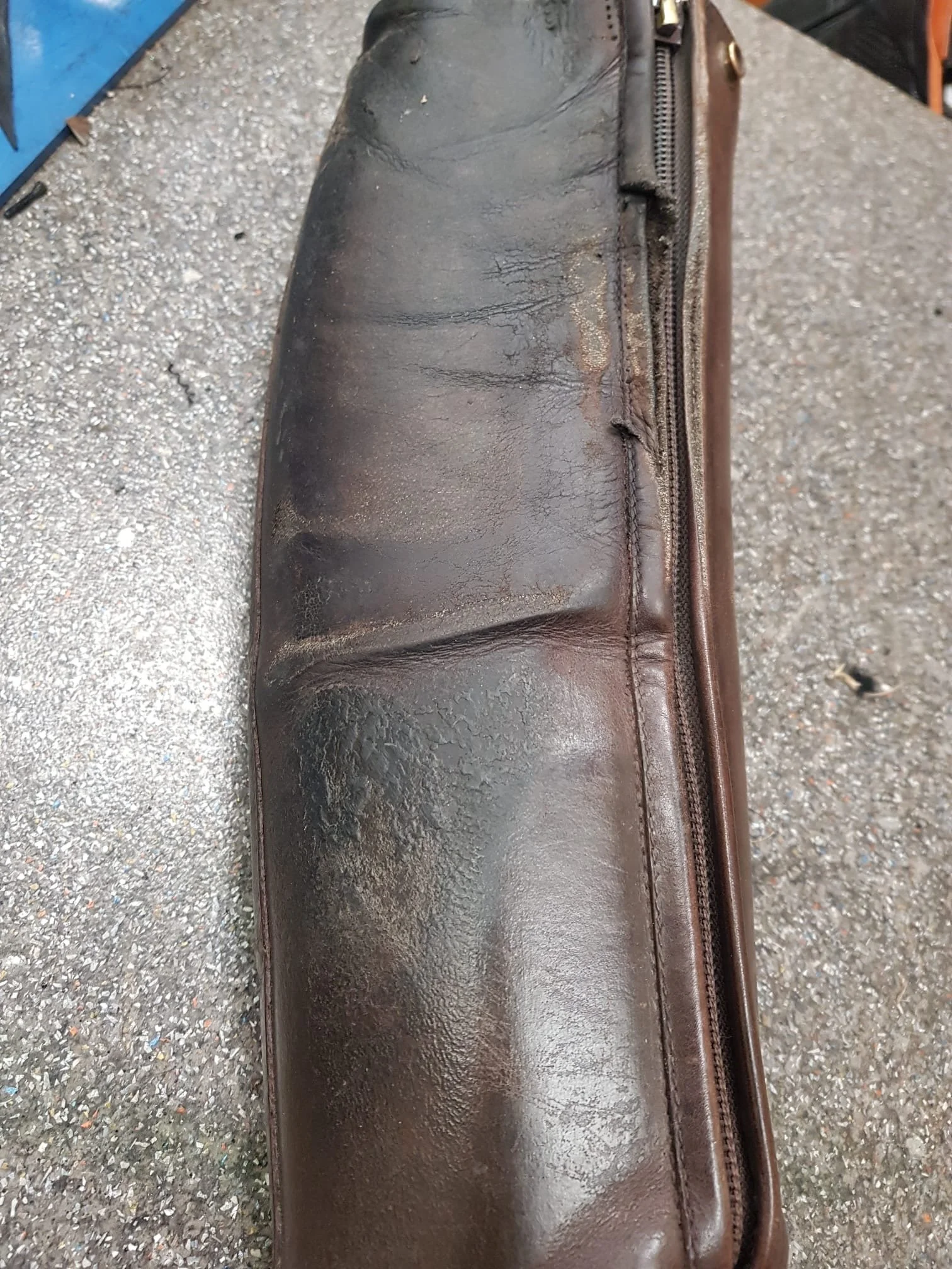 Ein altes, abgenutztes braunes Ledertäschchen mit Reißverschluss, liegt auf einem grauen, sprenkeligen Boden.