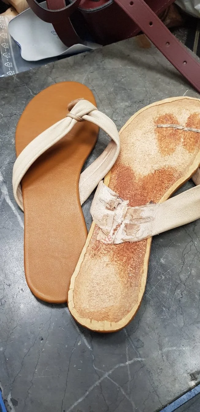Ein beschädigtes Paar Sandalen mit beige Bändern, das auf einer unebenem dunklen Oberfläche liegt.