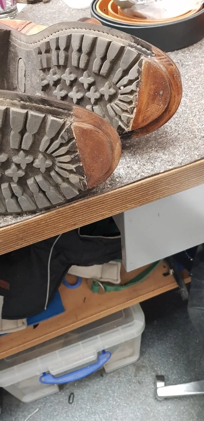 Nahaufnahme eines alten Ledersohlenpaars mit abgelaufenen Profilen, liegend auf einem Holztisch, im Hintergrund eine Werkzeugkiste.