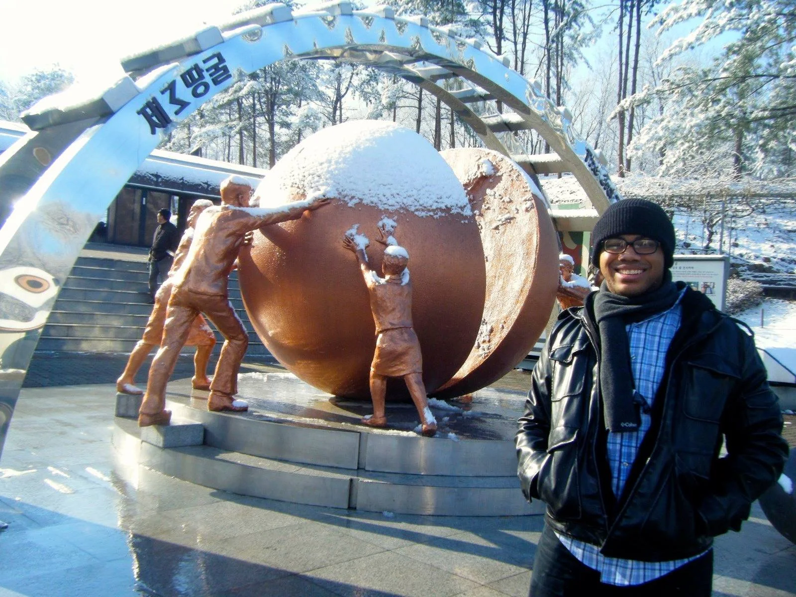 Aidan Hill wearing a black beanie, glasses, a black leather jacket, and a blue checkered shirt, smiling at the camera. In the background, there are snow-covered trees and a monument of statues representing the reunification of North & South Korea.