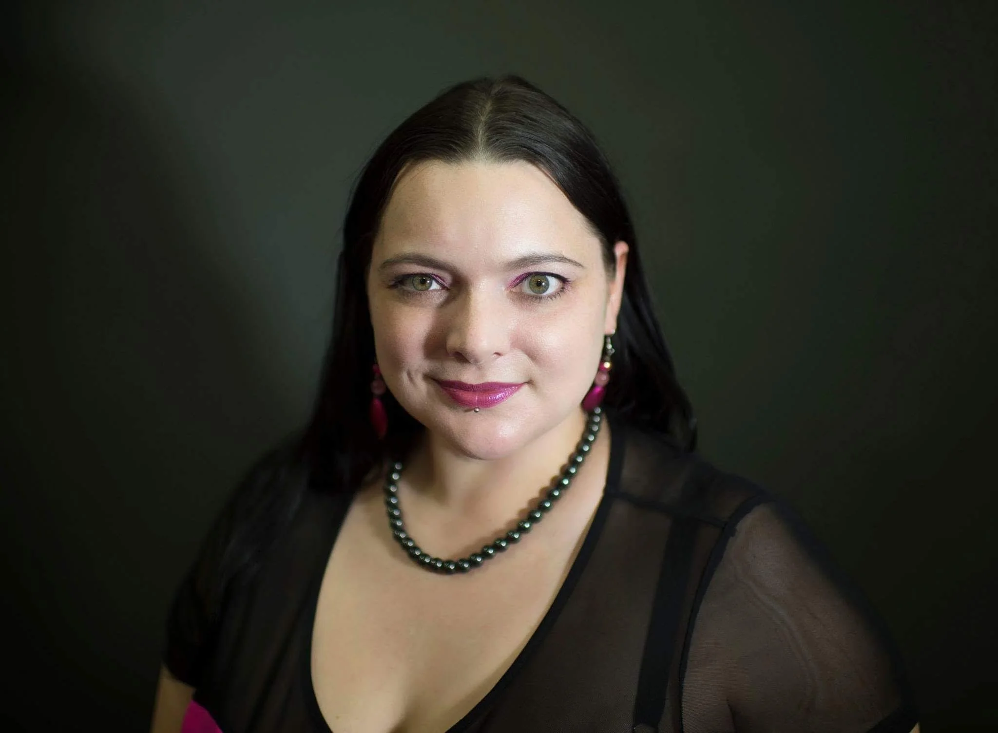 A woman with green eyes, dark hair, pink lipstick, and makeup, wearing a black top with sheer sleeves, a double strand pearl necklace, and pink earrings, smiling softly against a dark background.
