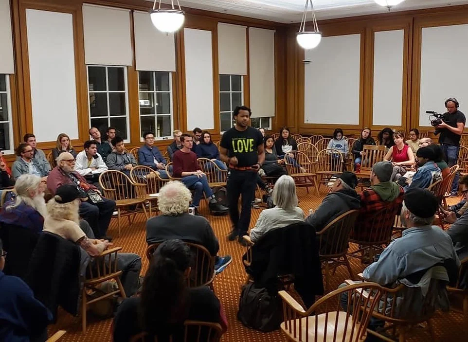 A diverse group of people seated in a circle in a well-lit room with wood-paneled walls, some listening attentively to Aidan Hill standing and speaking, and a cameraman recording the event.